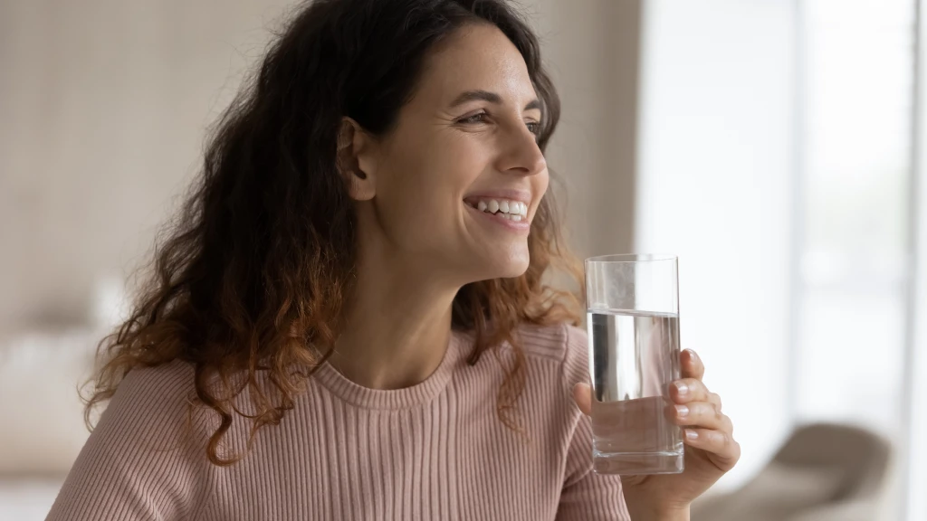 Mulher com blusa rosa claro sorrindo olhado para o lado enquanto se hidrata segurando copo d'água.